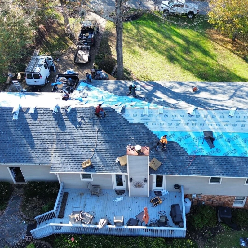 An aerial view of a house undergoing roof replacement, with workers installing blue underlayment and shingles.